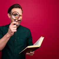 Photo of concentrated focused man looking at you through loupe using book to examine your knowledge isolated vivid color background